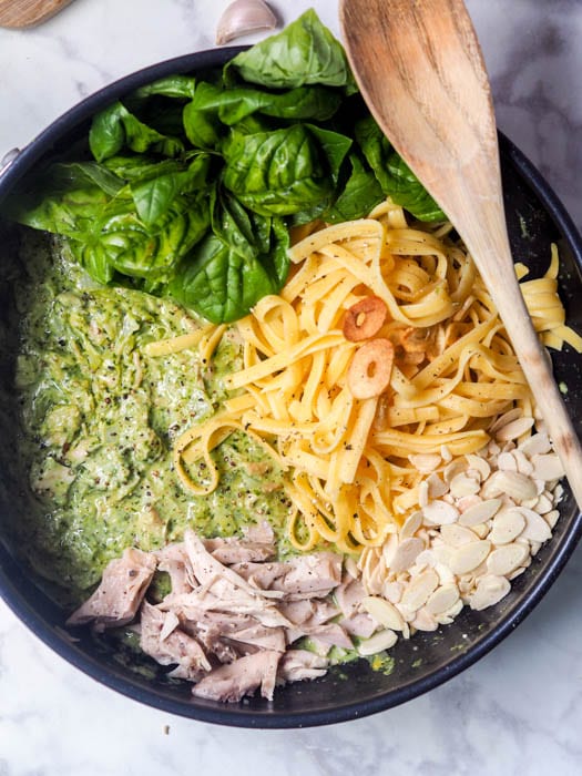Pesto chicken pasta being assembled in the pan with pasta, pesto, shredded chicken, sliced almonds and fresh basil