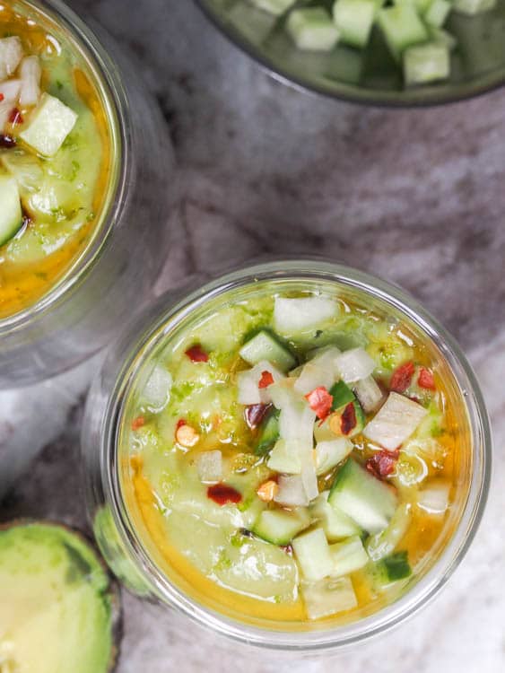 chilled avocado soup in small serving glasses topped with onions, cucumbers, oil and chili flakes
